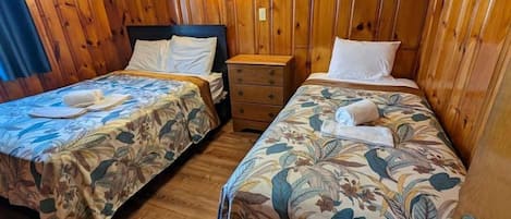 Une chambre avec deux lits, des murs en bois, une table de chevet et une lampe fixée au mur.