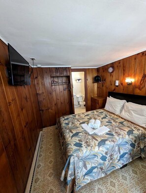Ein Schlafzimmer mit Holzwänden, einem Bett mit floralem Bettbezug, einem Nachttisch, einem an der Wand befestigten Fernseher und einer Uhr.