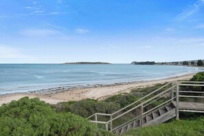 A coastal view with a wooden staircase leading down to a sandy beach, a clear sky, and a distant island.