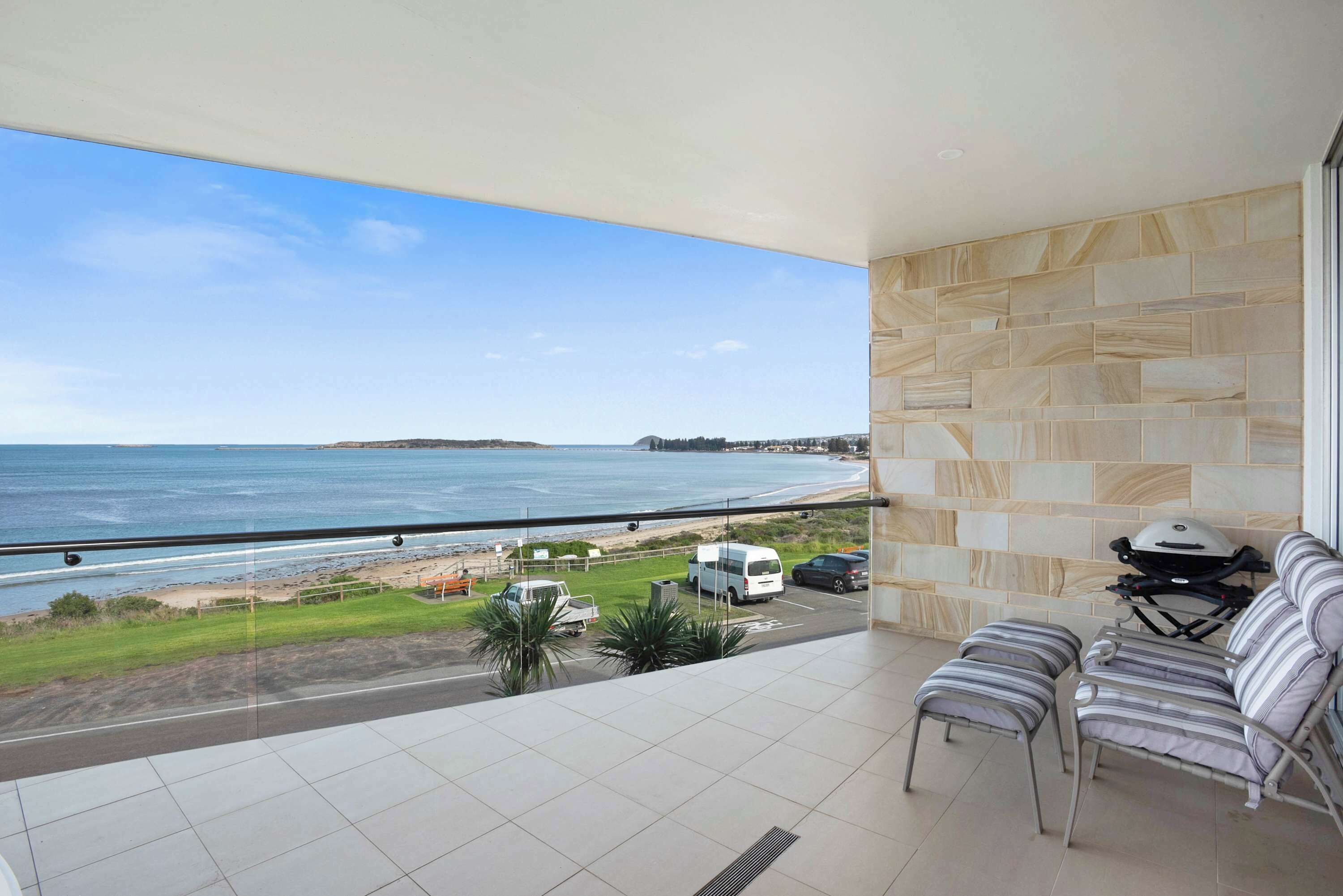 A balcony with a view of the sea and beach, a seating area with a grill, and a tiled floor.
