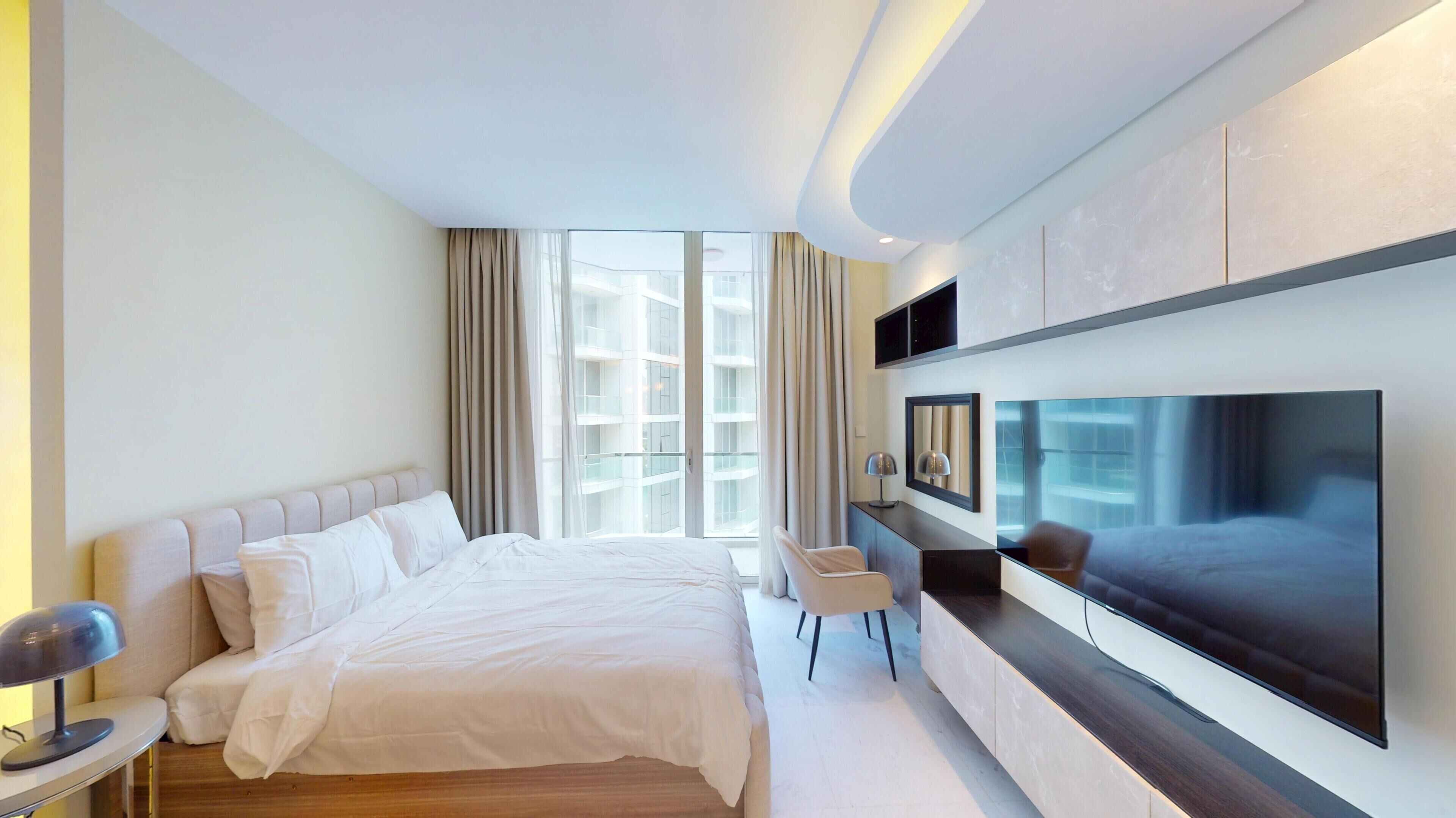 Quarto de hotel moderno com uma cama grande, televisão fixada na parede, escrivaninha com cadeira e janela com cortinas.