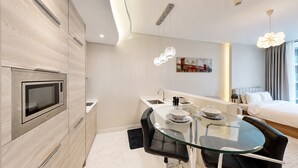 Moderni hotellihuone, jossa on pieni keittiö, lasinen ruokapöytä, sänky ja taulu seinällä.