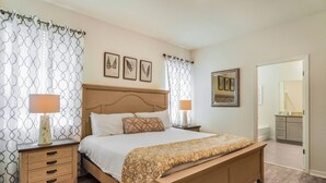 Une chambre à coucher comprenant un lit, deux tables de chevet, une commode, une fenêtre avec des rideaux à motifs et une porte donnant sur une salle de bain.