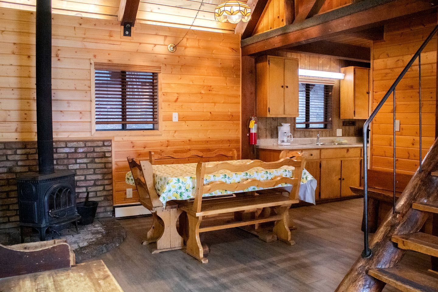 A cozy cabin interior with a wooden bench, a fireplace, a kitchen area, and a staircase.