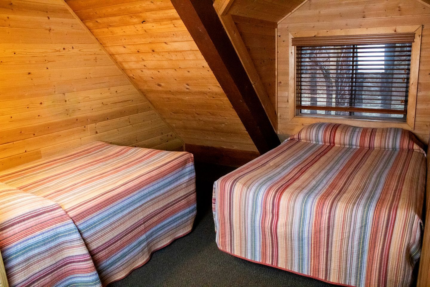 Une petite chambre mansardée, chaleureuse et accueillante, avec deux lits, une fenêtre avec des stores et des poutres en bois.