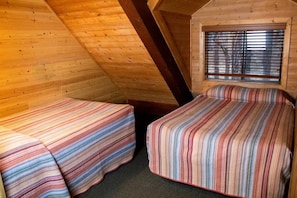Quarto de sótão pequeno e acolhedor, com duas camas, uma janela com persianas e vigas de madeira.
