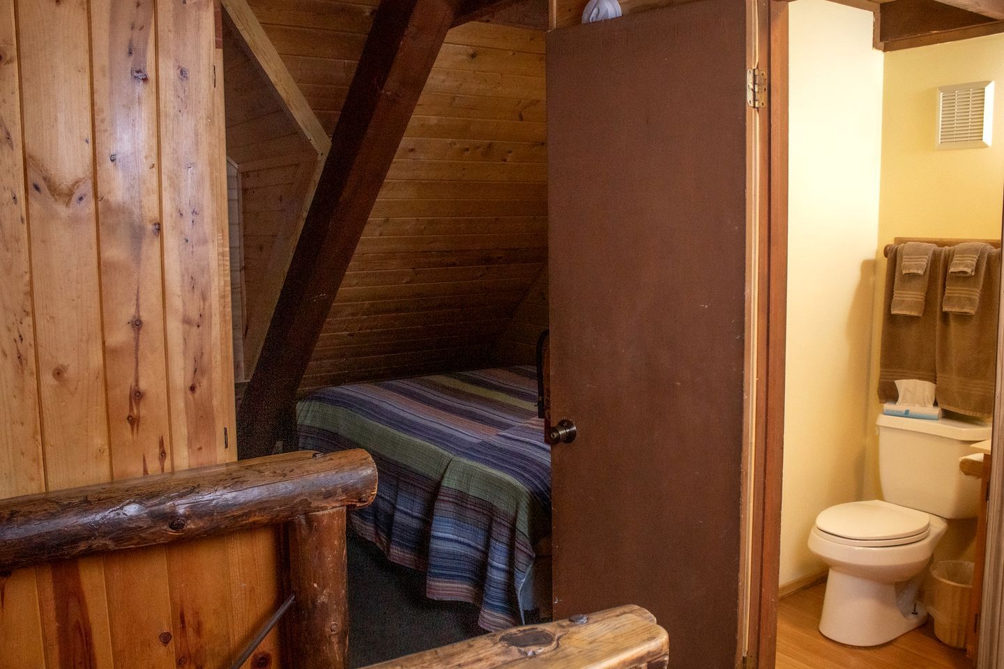 Un interior de madera con techo inclinado, una cama con manta a rayas y un inodoro blanco.