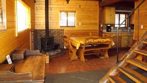 Interior de una cabaña de madera con mesa de comedor, bancos, chimenea y zona de cocina.