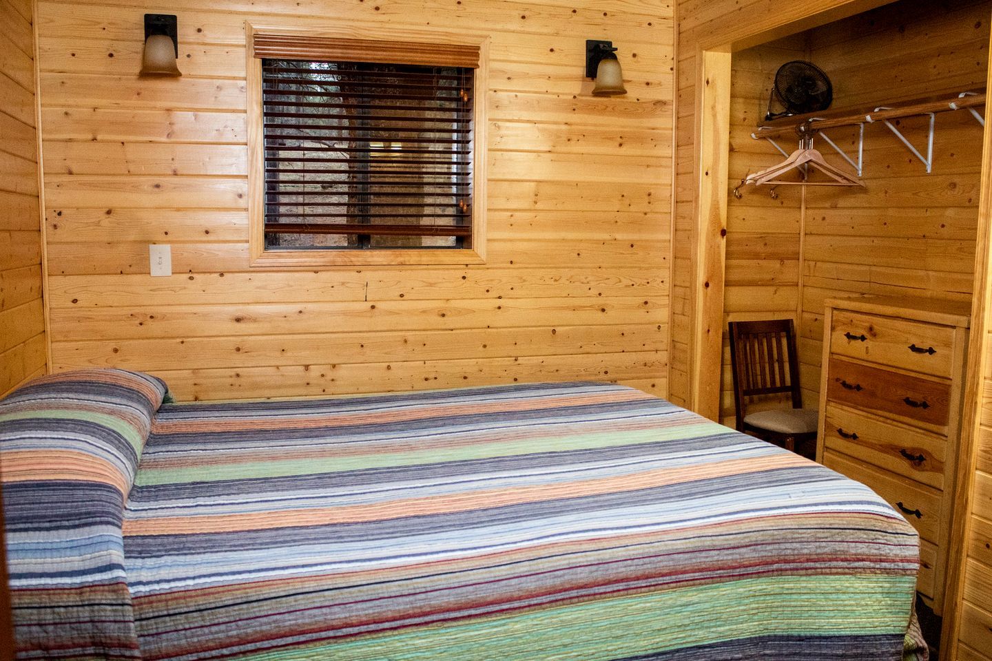 A wooden cabin room with a bed, a window with blinds, a dresser, and a hanging coat rack.