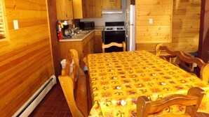 Interior de una cabaña de madera con mesa de comedor, sillas, zona de cocina y chimenea.