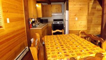A wooden cabin interior with a dining table, chairs, a kitchen area, and a fireplace.
