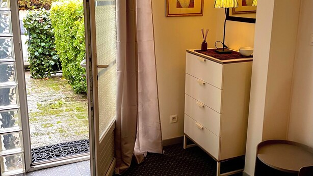 Quarto de hotel com porta de correr de vidro para um jardim, penteadeira branca com abajur e um quadro pendurado na parede.