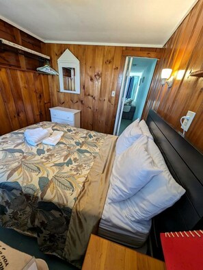 Une chambre avec des murs en bois, un lit avec une literie à motifs floraux, une table de chevet et une commode, ainsi qu’un miroir.