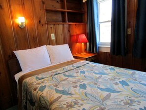A bed with a floral bedspread, a nightstand with a lamp, wooden walls, and a window with curtains. - Timeless Oceanfront Cottage Steps to Shore in North Truro, Massachusetts (North Truro)