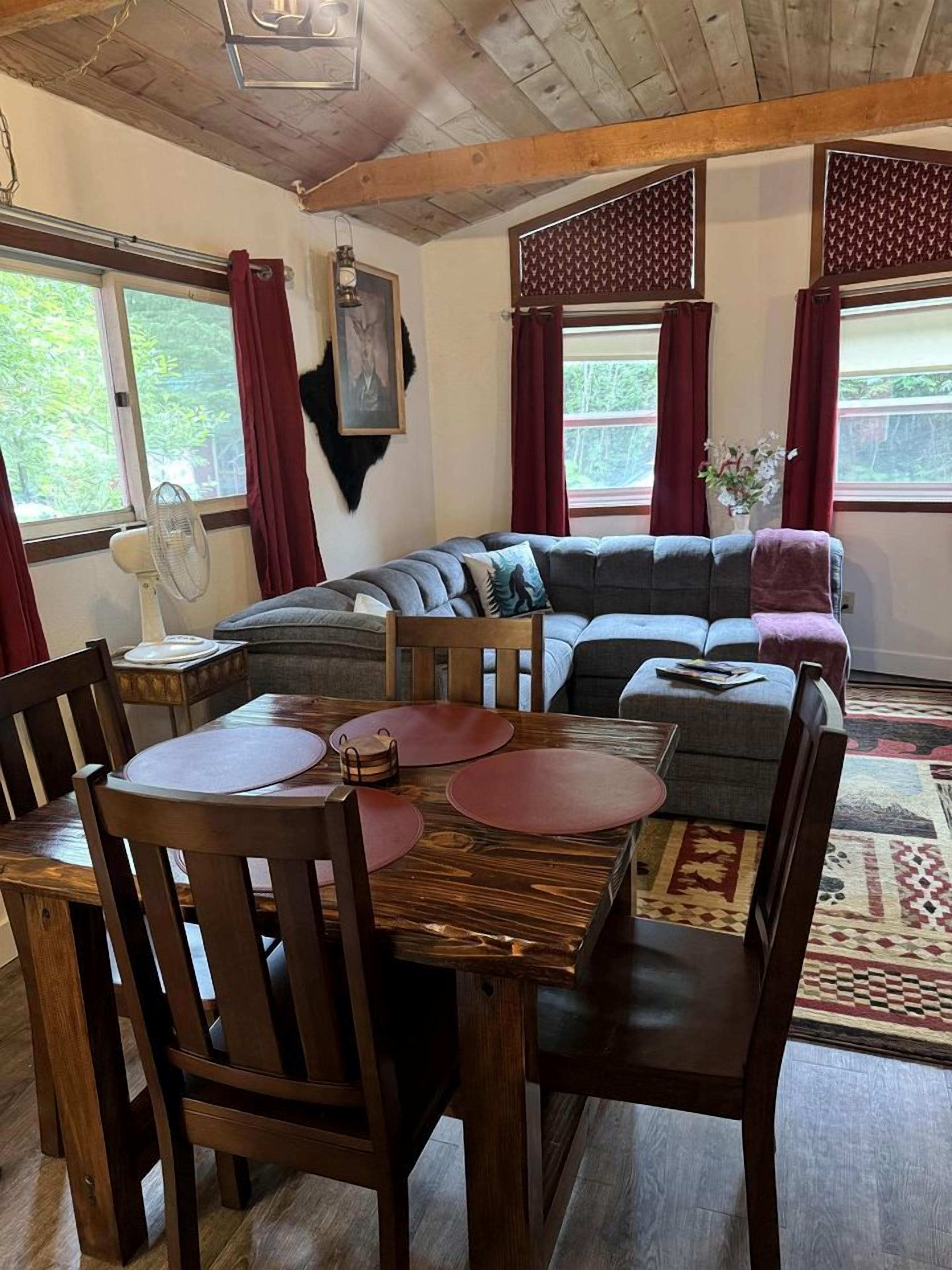 Una sala de estar acogedora con techo de madera, un sofá seccional gris, una mesa de comedor con sillas y un gran ventanal con cortinas.