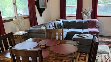Una sala de estar acogedora con techo de madera, un sofá seccional gris, una mesa de comedor con sillas y un gran ventanal con cortinas.