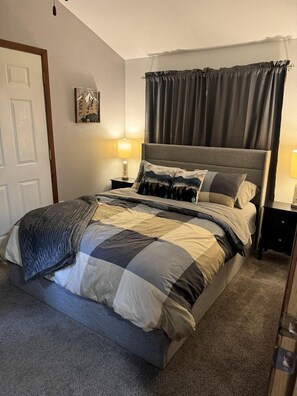 Un letto ben rifatto con copripiumino grigio e bianco a quadretti, una coperta grigia e una testiera con tenda. C'è un comodino con una lampada e un piccolo oggetto decorativo.