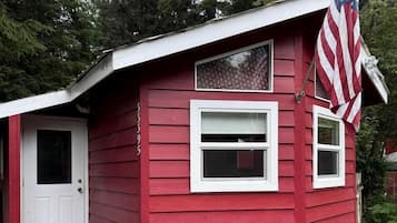 Una cabaña de madera roja con una puerta blanca, un pequeño porche y una bandera estadounidense.