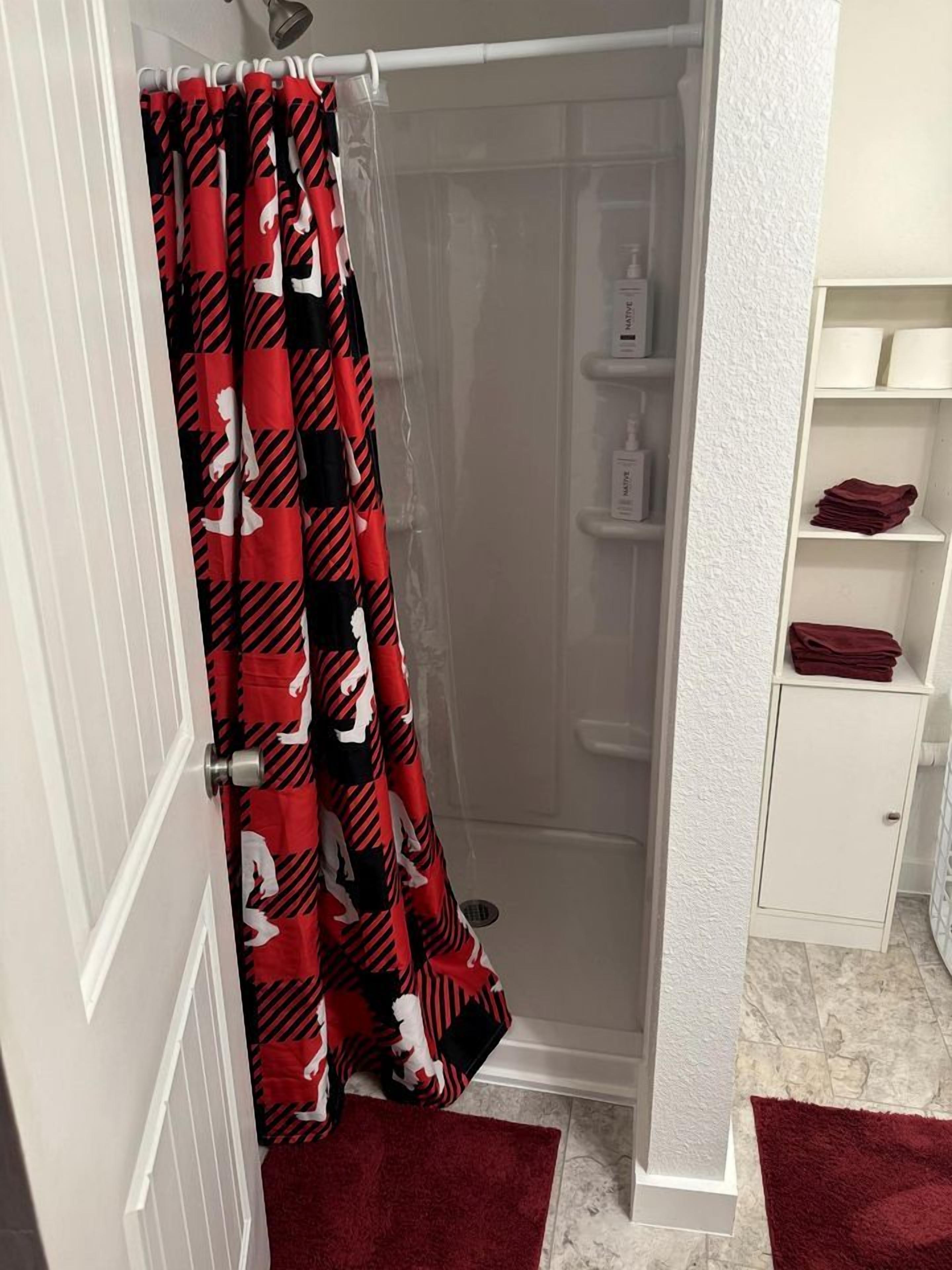 A bathroom with a shower, a white door, a white shelf with toiletries, and a red towel on the floor.