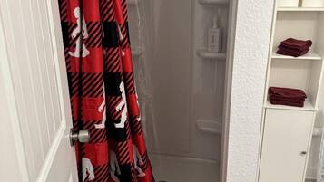A bathroom with a shower, a white door, a white shelf with toiletries, and a red towel on the floor.