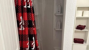 Ett badrum med dusch, en vit dörr, en vit hylla med toalettartiklar och en röd handduk på golvet.