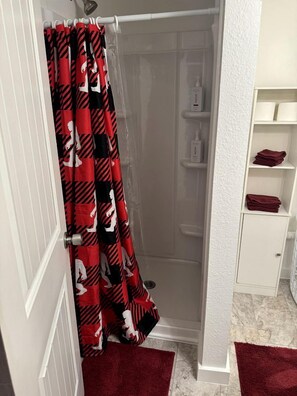 Bagno con doccia, una porta bianca, uno scaffale bianco con articoli da toilette e un asciugamano rosso sul pavimento.