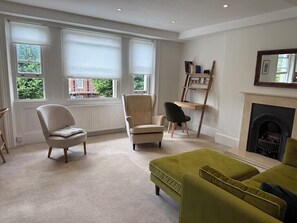 A room with a fireplace, a green sofa, a beige armchair, and a wooden bookshelf. - Light, airy, great location, newly decorated and furnished. Great views. (London)