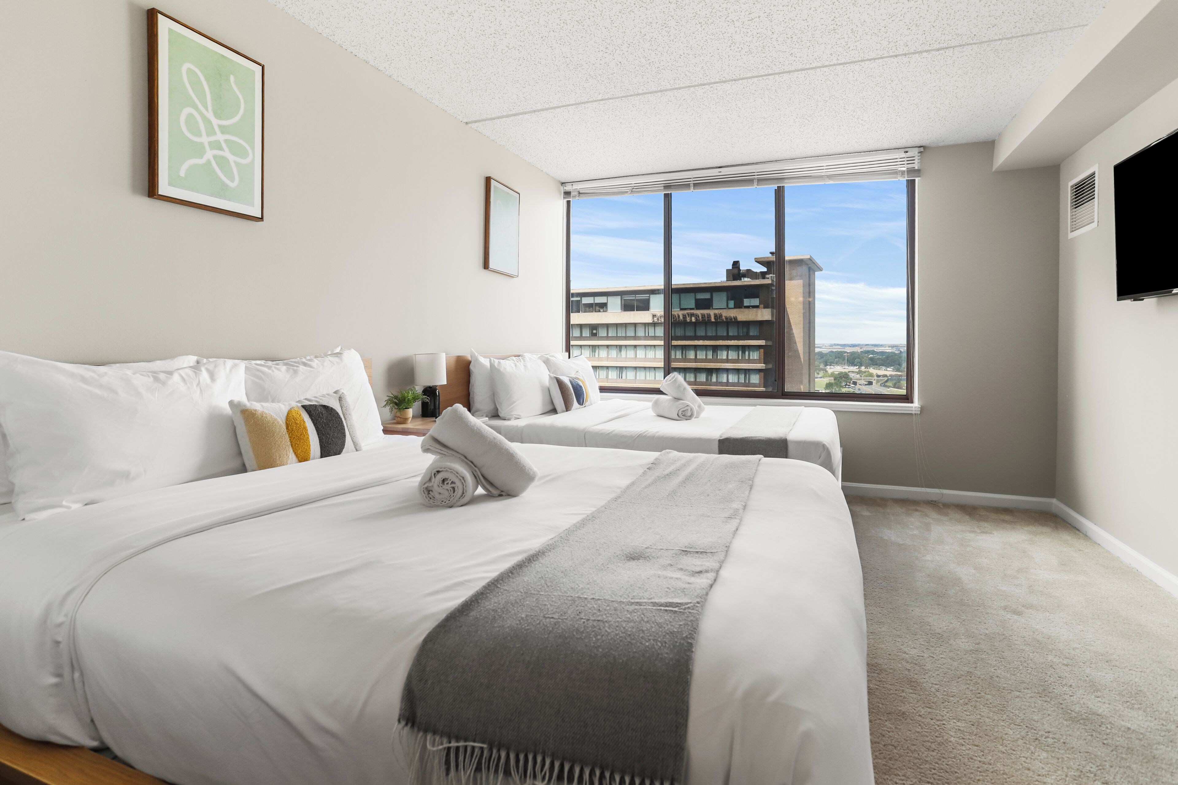 A hotel room with two beds, a large window with a city view, a flat-screen TV, and a ceiling with a speckled pattern.