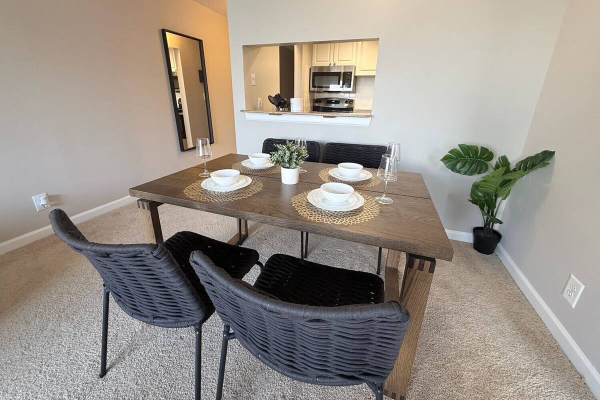 A dining area with a wooden table, four chairs, a potted plant, and a kitchen in the background.
