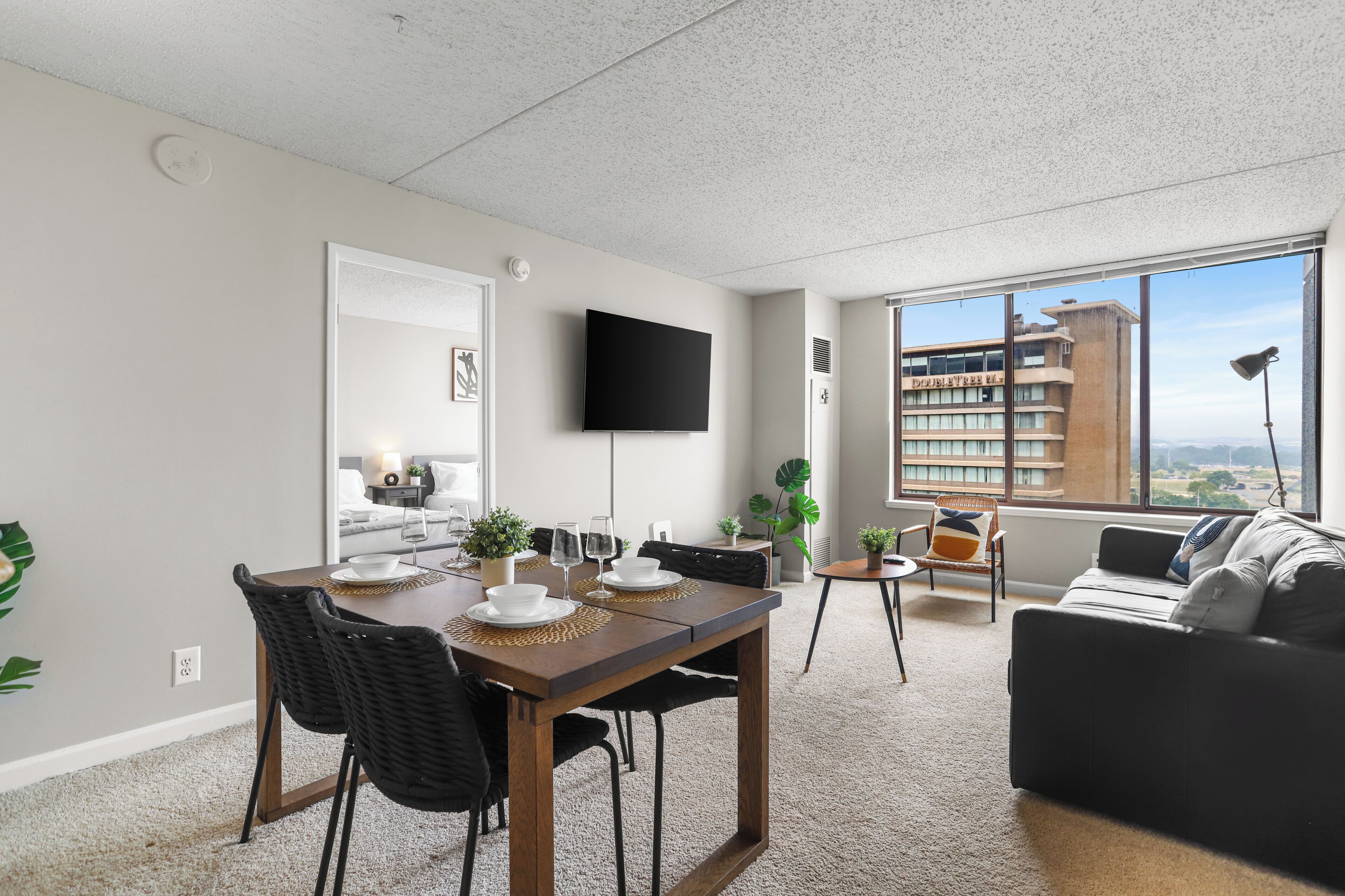 Una sala de estar moderna con comedor, un televisor de pantalla plana montado en la pared y un amplio ventanal con vistas a un edificio y paisaje.