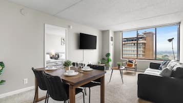 Un salon moderne comprenant un coin repas, un téléviseur à écran plat fixé au mur et une grande fenêtre donnant sur un bâtiment et un paysage.