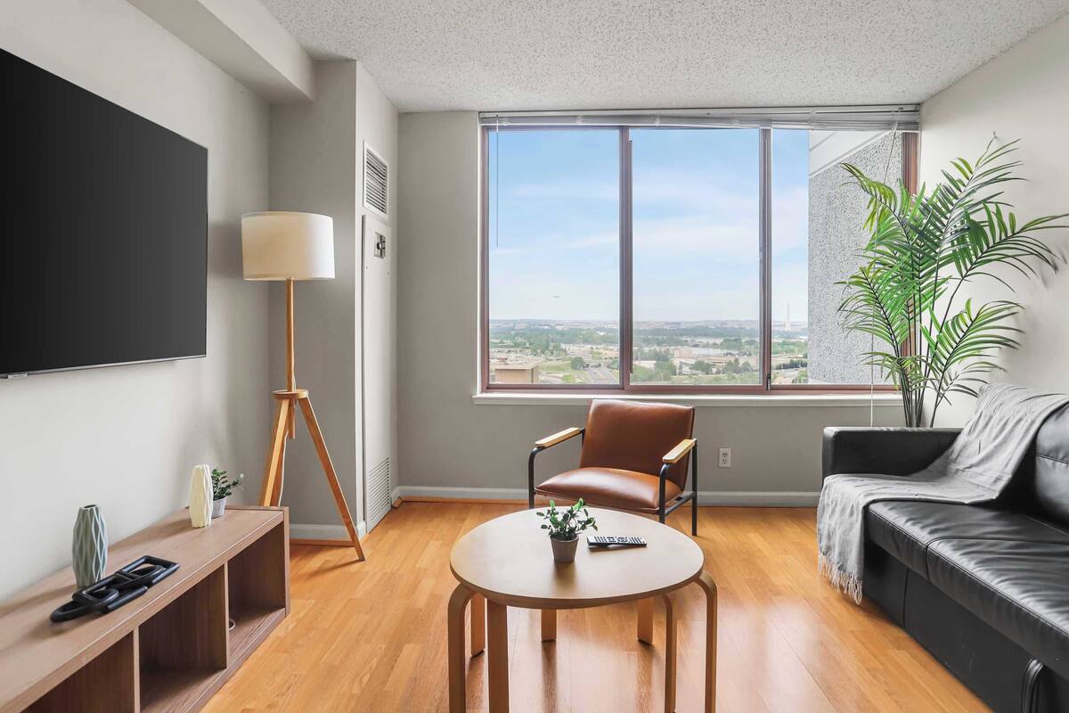 A modern living room with a large window, a flat-screen TV, a brown leather chair, a black leather sofa, and a round wooden table with a plant.