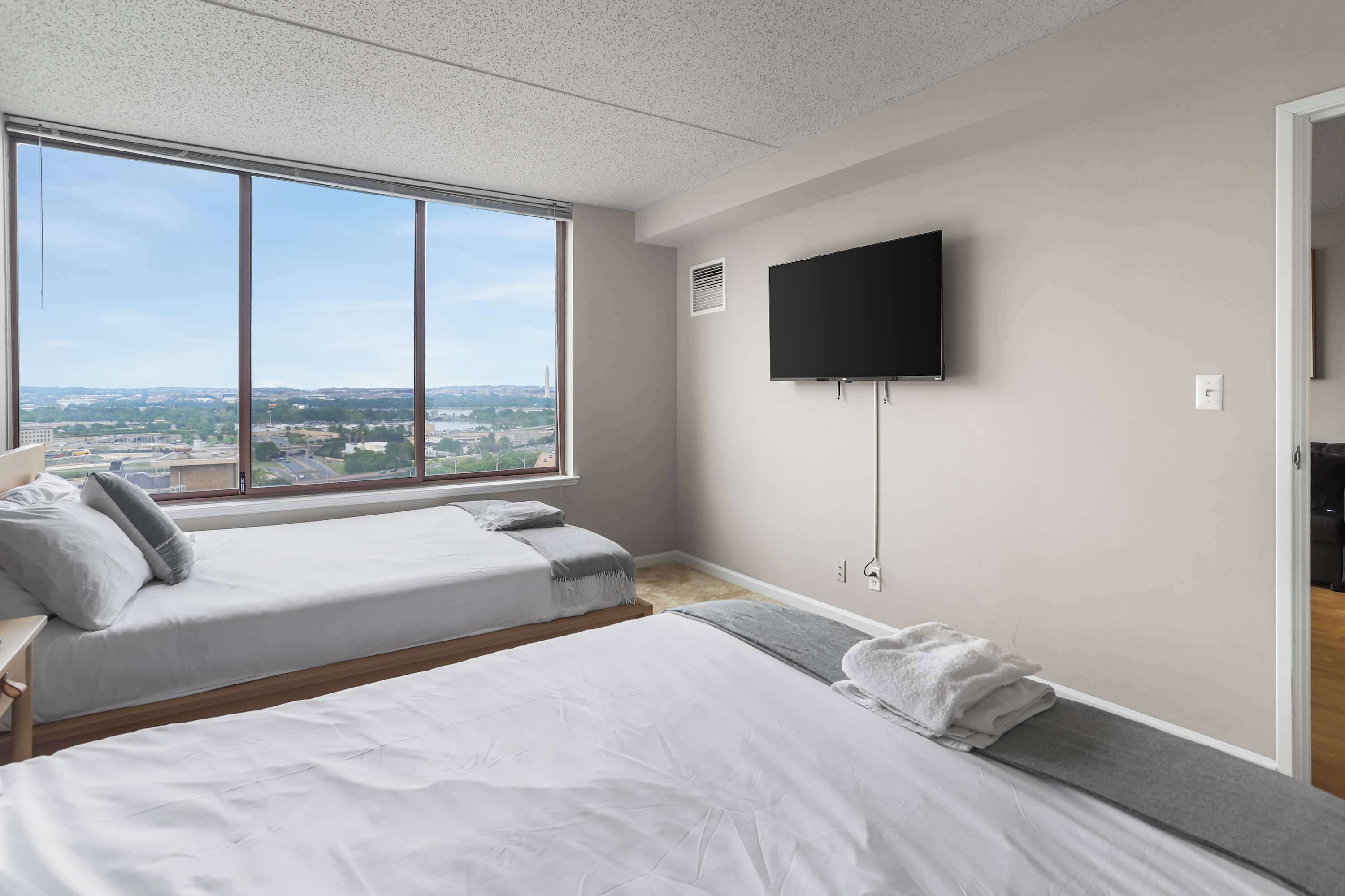 A modern hotel room with two beds, a large window, a flat-screen TV, and a city view.