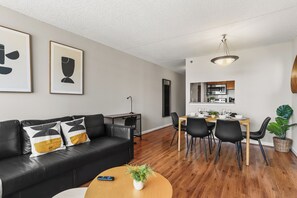 Una sala de estar moderna con un sofá de cuero negro, una mesa de comedor con sillas y una cocina visible a través de un vano abierto.