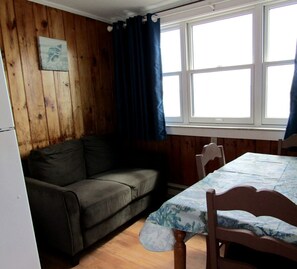 A room with wooden walls, a sofa, a table with a patterned tablecloth, and a window with curtains. - Seaside Escape with Coastal Vibes in North Truro, Massachusetts (North Truro)
