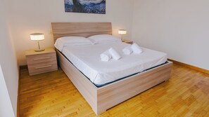 A neatly made bed with white linens and towels, a wooden headboard, bedside tables with lamps, and a wooden floor. - Expo Smart Living - Two Bedroom Apartment, Sleeps 6 (Verona)