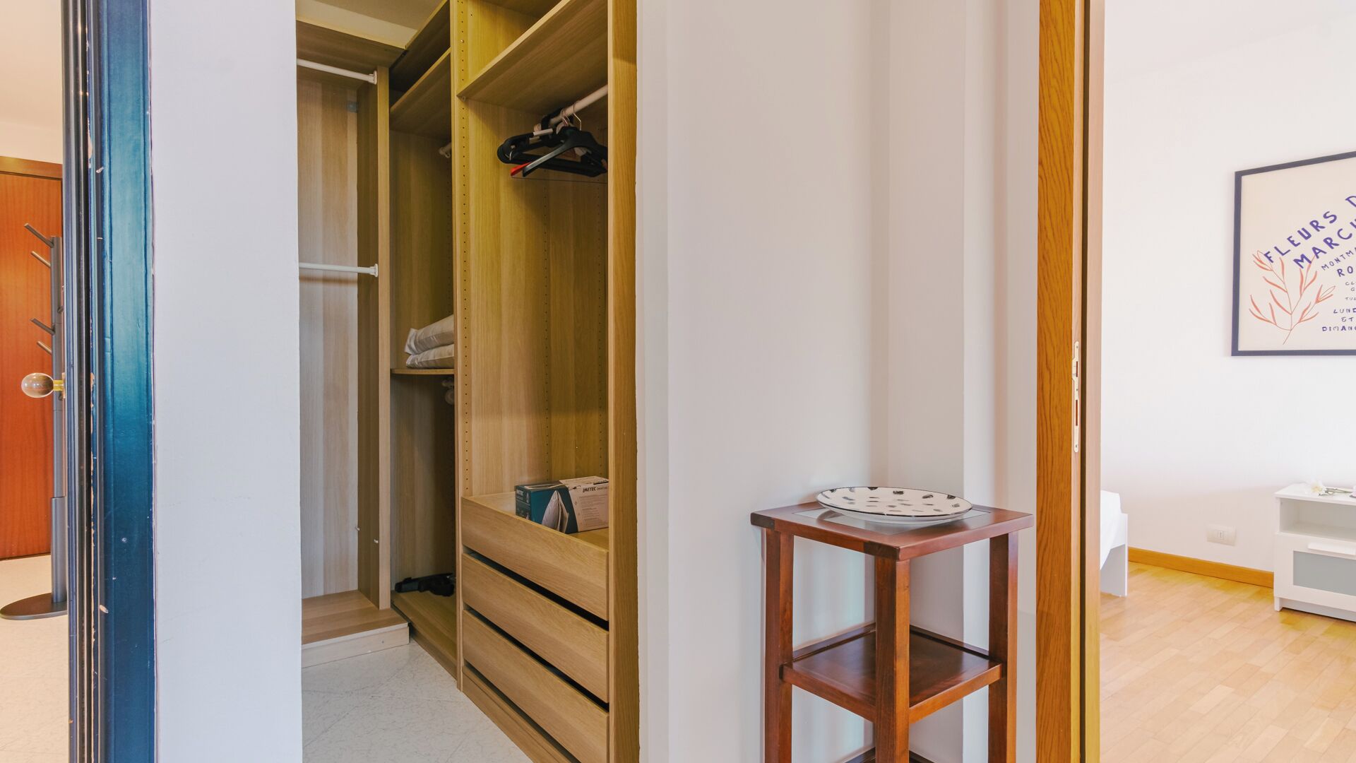 A room with a wooden wardrobe, a small table with a plate, and a framed picture on the wall.