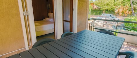 A balcony with a table and chairs, a bed in the adjacent room, and a view of parked cars and greenery.