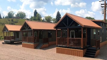 A row of cabins with wooden decks and metal roofs, set on a gravel ground with trees in the background.
