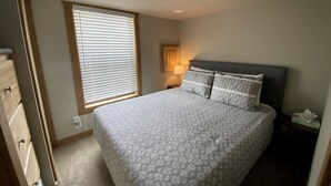 A bedroom with a bed, a nightstand, a lamp, a window with blinds, and a door.