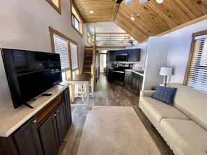A modern interior with a flat-screen TV, a sofa, a kitchen area, and a staircase leading to an upper level.