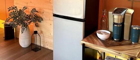 A compact kitchen with a refrigerator, an air conditioner, a coffee maker, and a small table with a plant and a sign.