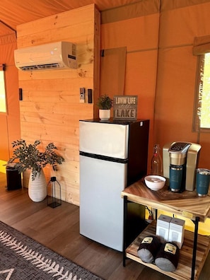 A compact kitchen with a refrigerator, an air conditioner, a coffee maker, and a small table with a plant and a sign.