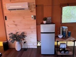 Cocina compacta con aire acondicionado integrado, refrigerador, pared de madera, una mesa pequeña con cafetera y una planta en maceta.