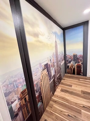 A room with a large cityscape mural, wooden flooring, and a black-framed window. - Deco Studio Retreat: Fireplace, WiFi, Queen Bed (Reno)