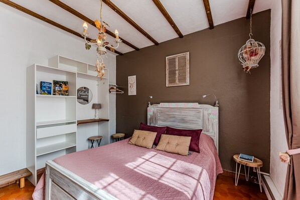 A bedroom with a bed, a white wardrobe, a small table, a hanging light fixture, and a window with blinds.