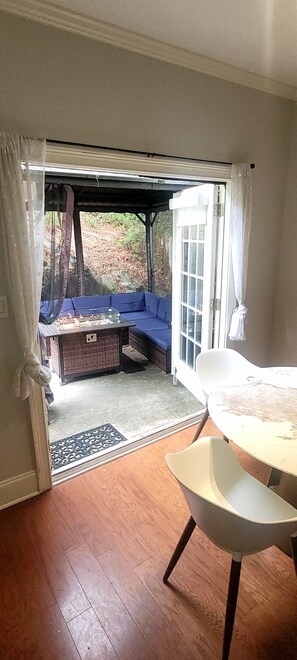 Ein Zimmer mit Holzboden, einem weißen Stuhl, einem Tisch und Blick durch eine Glasdoor auf einen Außenbereich mit blauer Couch und Grill.