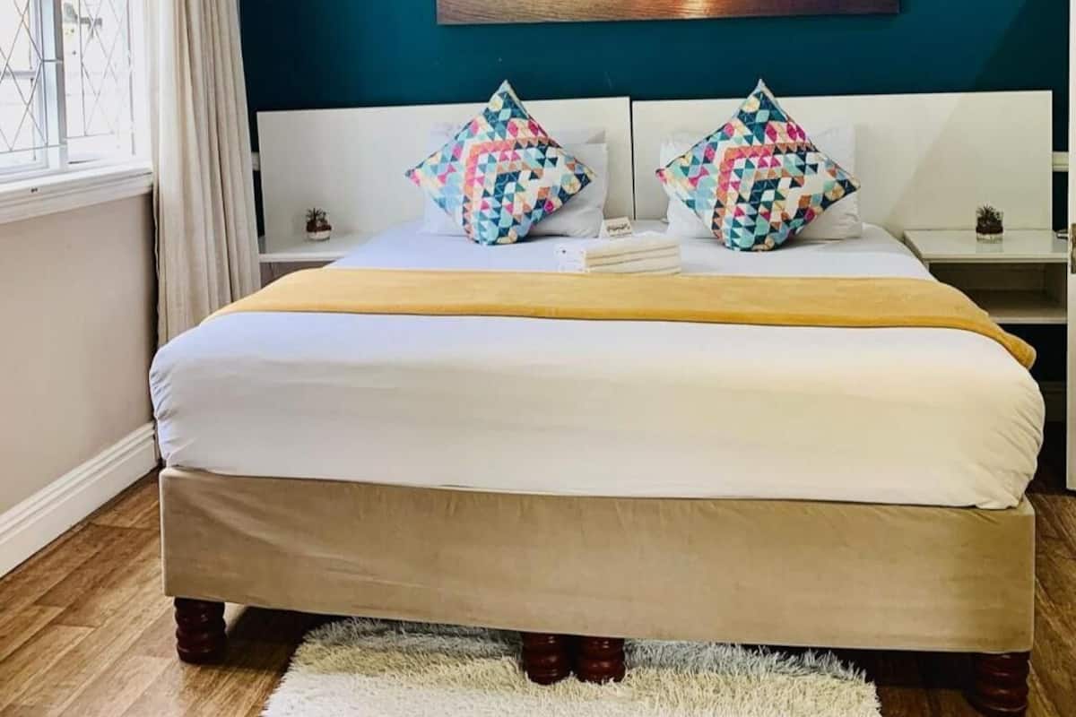 A bed with a white mattress and a yellow blanket, two patterned pillows, a small bedside table with a plant, and a painting of a sunset on the wall.
