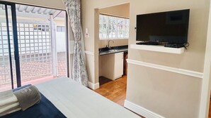 Quarto de hotel com uma cama, televisão fixada na parede, pequeno espaço de cozinha e porta de vidro deslizante para uma área externa.