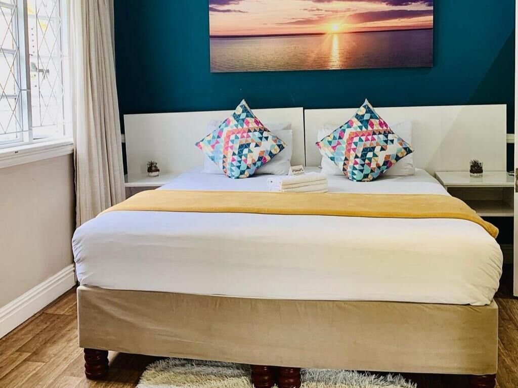 A double bed with a yellow throw blanket, two patterned pillows, a white headboard, and a sunset painting on the wall.